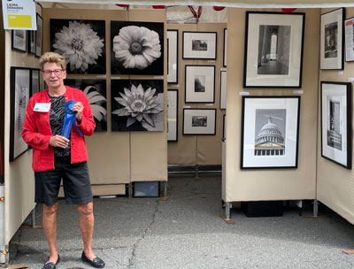 Laura Denardo stands in her booth surrounded by black and white photography holding her blue ribbon for Best Photography at Tephra ICA Arts Festival